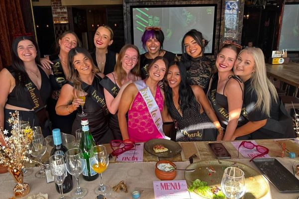 A big hens group smiles around a long restaurant table, the bride in a sash
