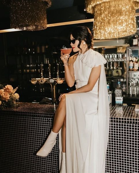 A woman in a white dress and sunglasses sipping a cocktail at a glamorous bar