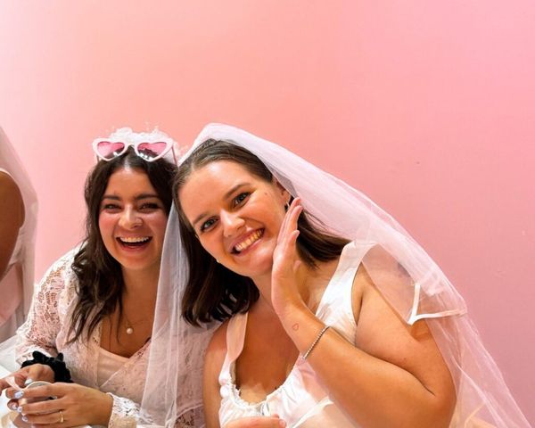 The bride in her veil waving and smiling with her friend in heart sunglasses at the class table