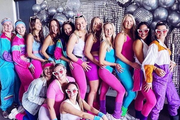 Hens group in bright retro tracksuits and heart sunglasses in front of silver balloons