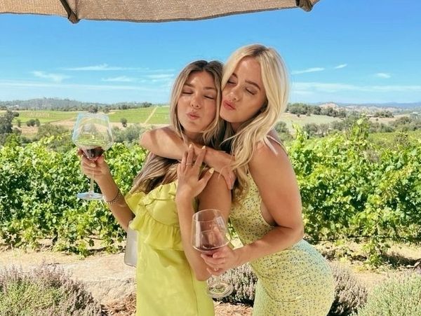 Two friends in summer dresses hugging among the vines with wine in hand