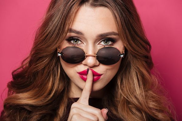 A woman in round sunglasses and red lipstick holding a finger to her lips