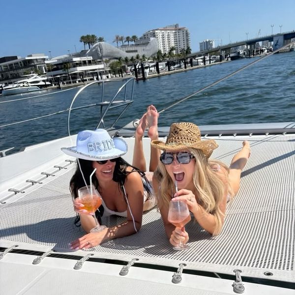 Two friends in cowboy hats lying on a catamaran deck with drinks, waterfront apartments behind