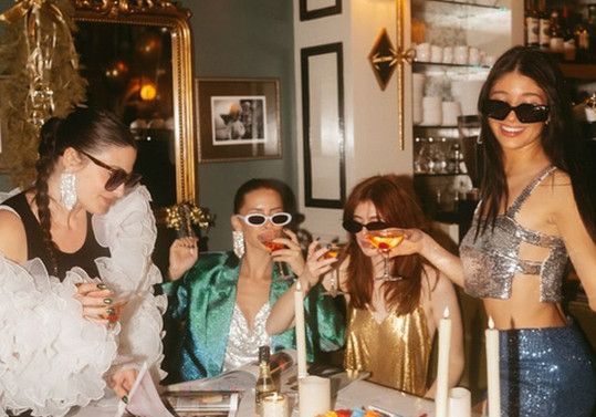 Friends in sunglasses, sequins and ruffles raising cocktails at a candlelit table