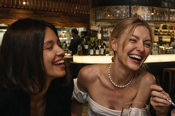 Two friends laugh over share plates and a pink cocktail at a restaurant