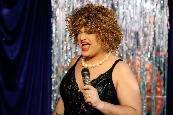 A drag queen in pearls and black sequins singing into a microphone in front of a tinsel curtain
