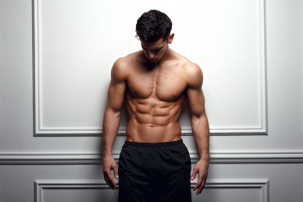 A shirtless man in black shorts leaning against a white panelled wall