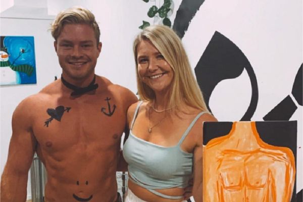 A shirtless waiter in a bow tie beside a guest holding her painting of him