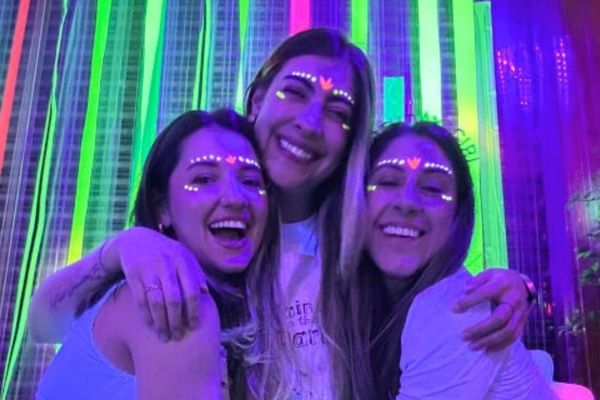 Three friends with glowing face paint hug and laugh under UV light at a paint class
