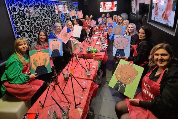 A hens group at a long table holding up paintings of a shirtless man