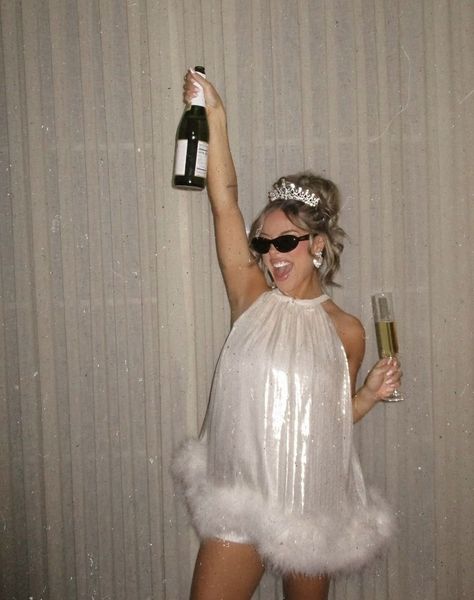 A bride to be in a tiara and sunglasses holding a bottle of bubbles up high