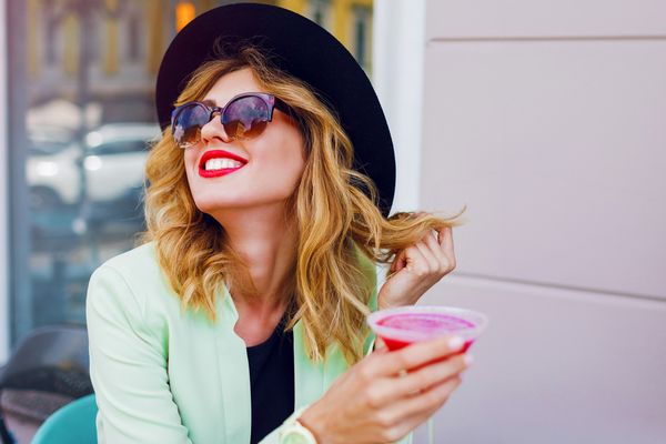 A woman in a hat and sunglasses smiling with a pink cocktail