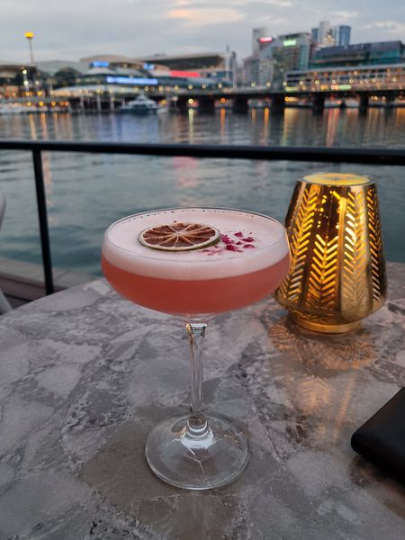 A pink cocktail on a waterfront table with the city skyline lit up across the water