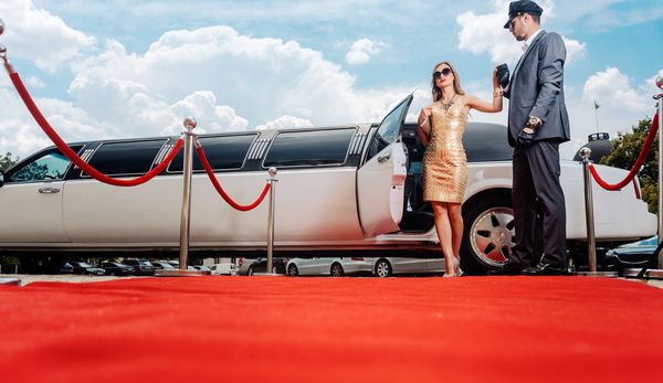 A chauffeur helping a woman in a gold dress out of a white limousine on a red carpet