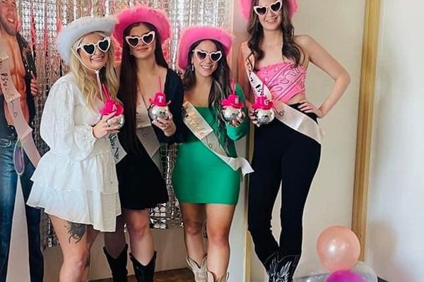 Four friends in pink cowgirl hats, sashes and boots pose by a tinsel backdrop