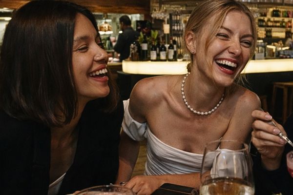 Two friends laughing together at a warmly lit restaurant table with a glass of white wine