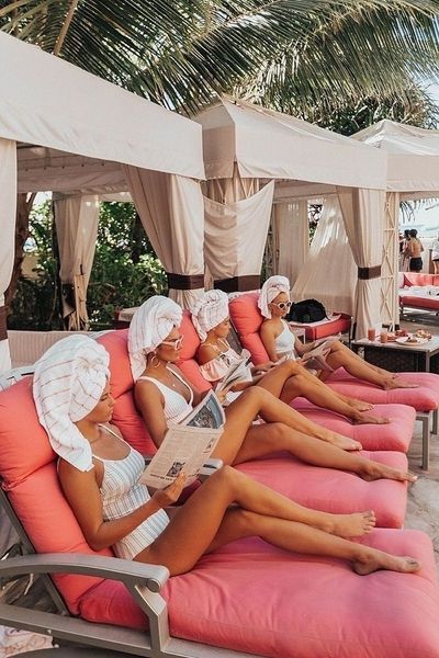 Friends in towel turbans and white swimwear reading on pink sun loungers under poolside cabanas