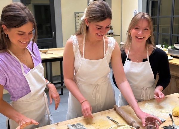 Three friends in aprons laughing as they shape gnocchi dough at a long timber bench