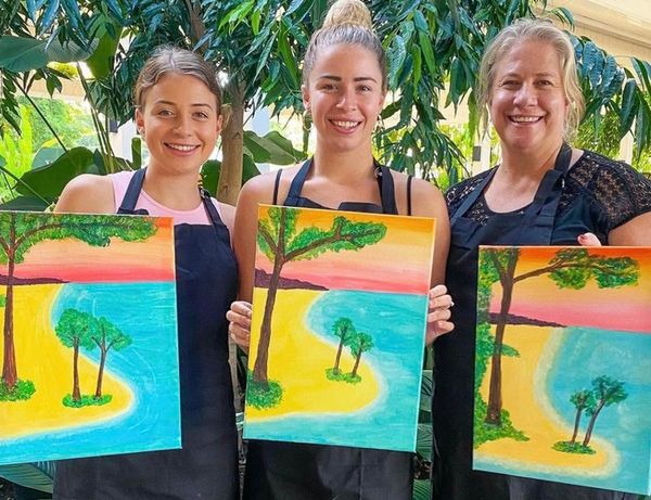 Three friends in aprons holding up their beach and tree paintings among the greenery