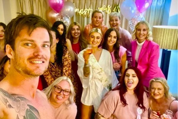 A shirtless performer taking a selfie with a hens group and pink balloons