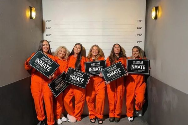 Six women in orange jumpsuits holding inmate signs at a mugshot height wall