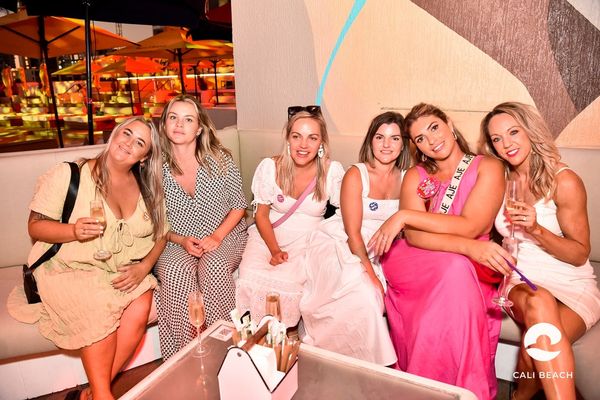 Hens group in summer dresses seated together on a white lounge, the bride wearing a sash