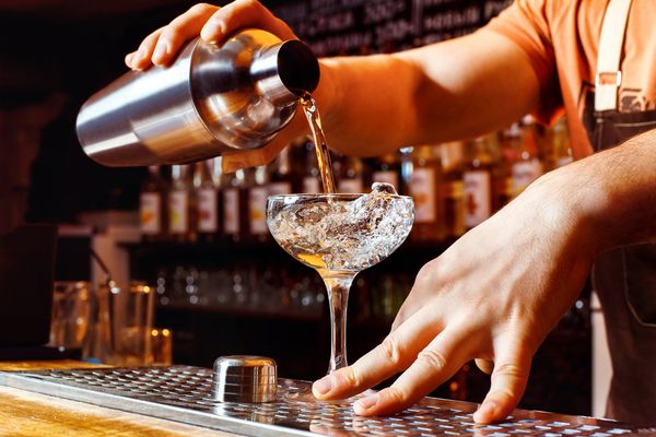 A bartender pours a cocktail from a shaker into a salt-rimmed coupe
