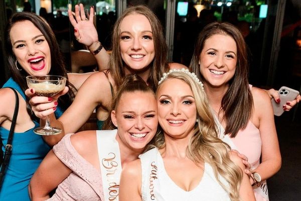 Bride and friends in sashes crowd together smiling, holding glasses at a night venue