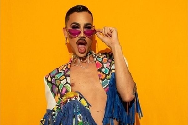 A drag queen in heart glasses and a fringed jacket striking a pose