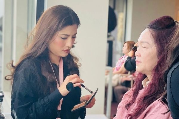 A makeup artist brushes on powder while a guest sits back in a director chair