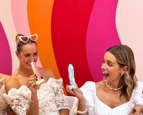 Two friends in white dresses laughing as they hold up their cheeky creations