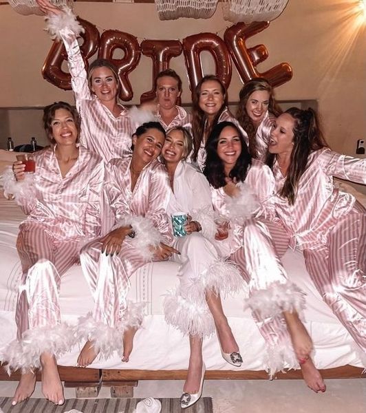 A hens group in pink satin pyjamas on a bed beneath Bride balloons