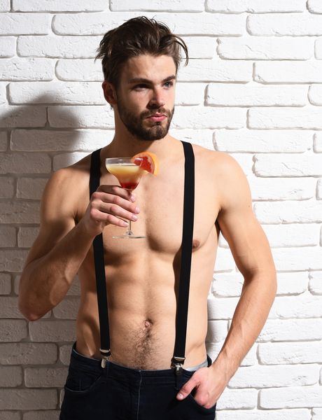 A shirtless waiter in braces holding a cocktail against a white brick wall