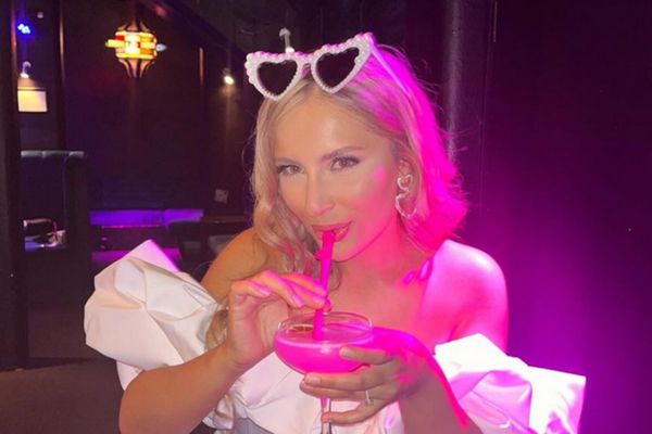 A hen in white with heart sunglasses sipping a pink cocktail under pink light