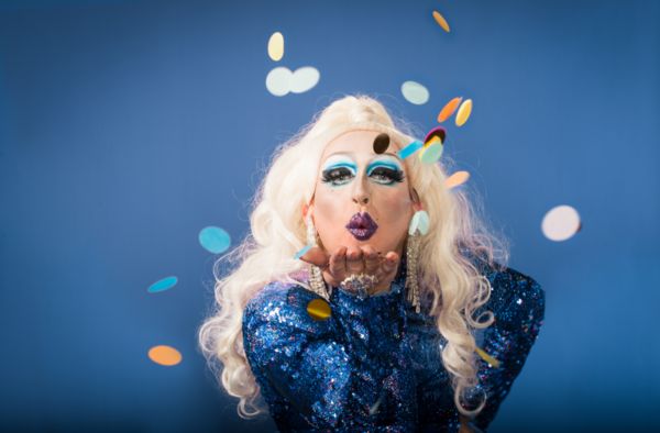 A drag queen in blue sequins blowing a kiss through falling confetti