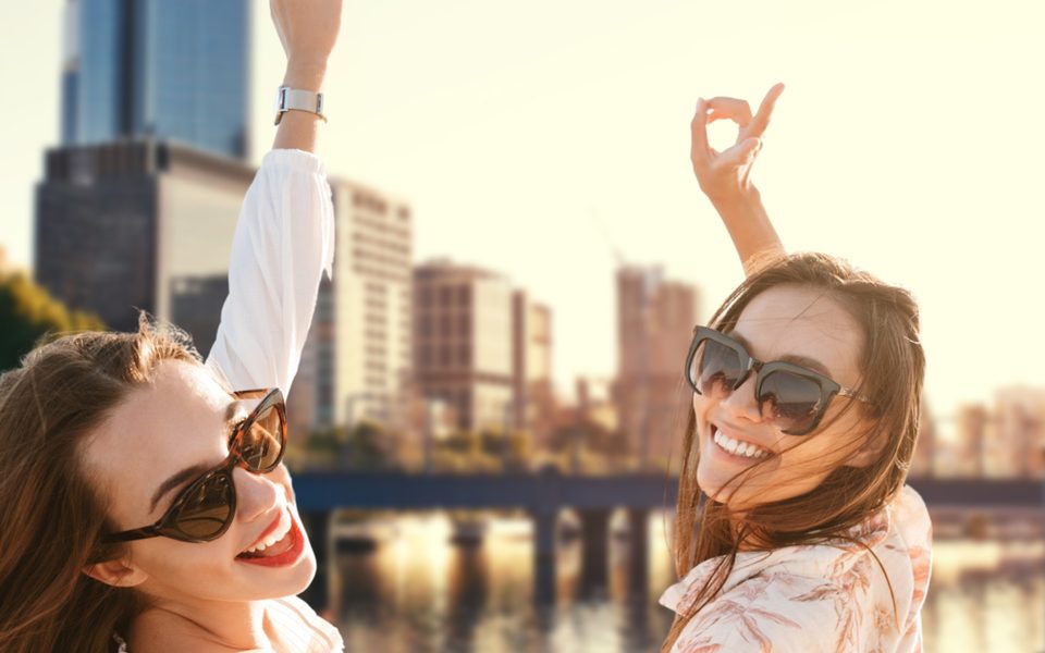 Two friends in sunglasses laughing by the river at sunset, the city towers behind them