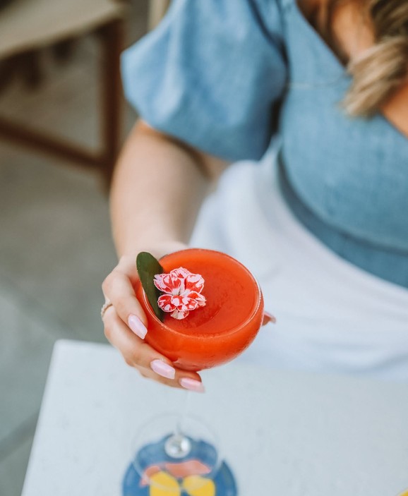 A hand holding out a coral drink with a pink and white flower in a coupe glass
