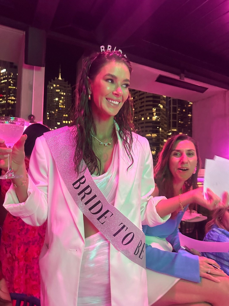 Britt in her Bride to Be sash and a white blazer, glass up, under pink lights at night