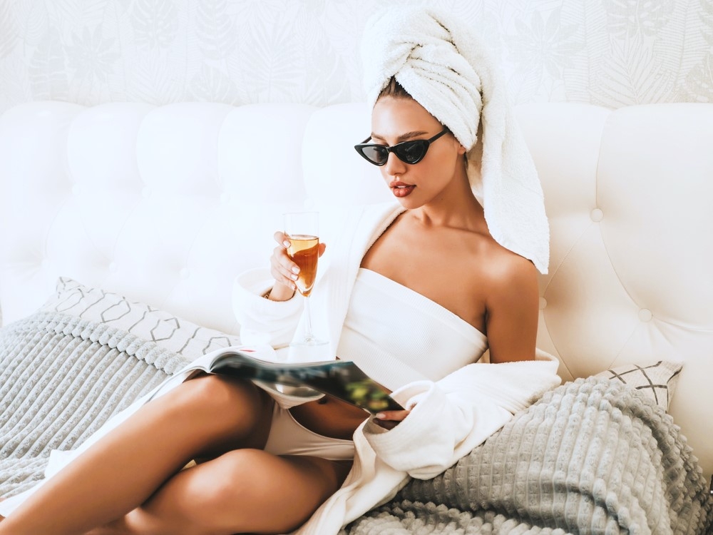A woman in a robe and towel turban reading a magazine on a white bed