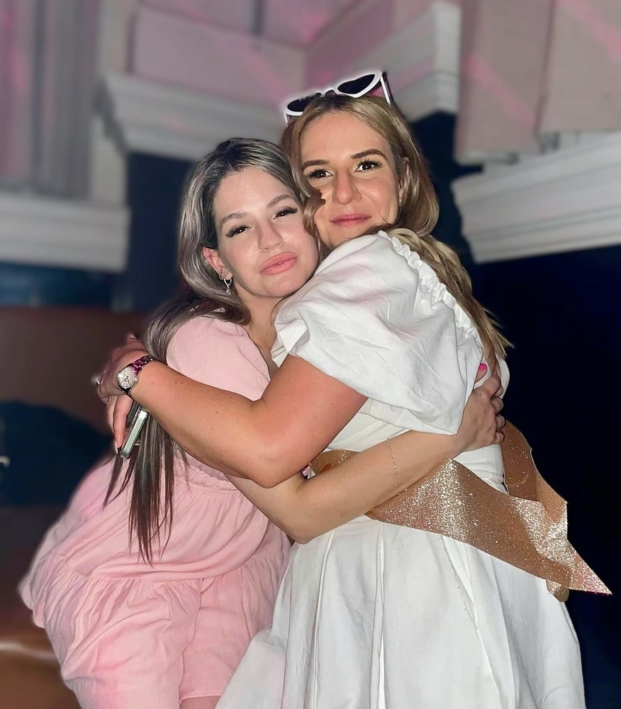 A bride to be in a white dress and gold sash hugging a friend in pink at her hens night