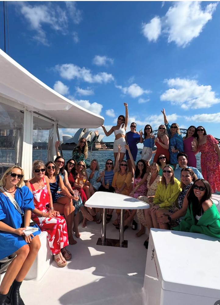 Britt's hens crew on the deck of the catamaran with their arms up, the harbour behind them