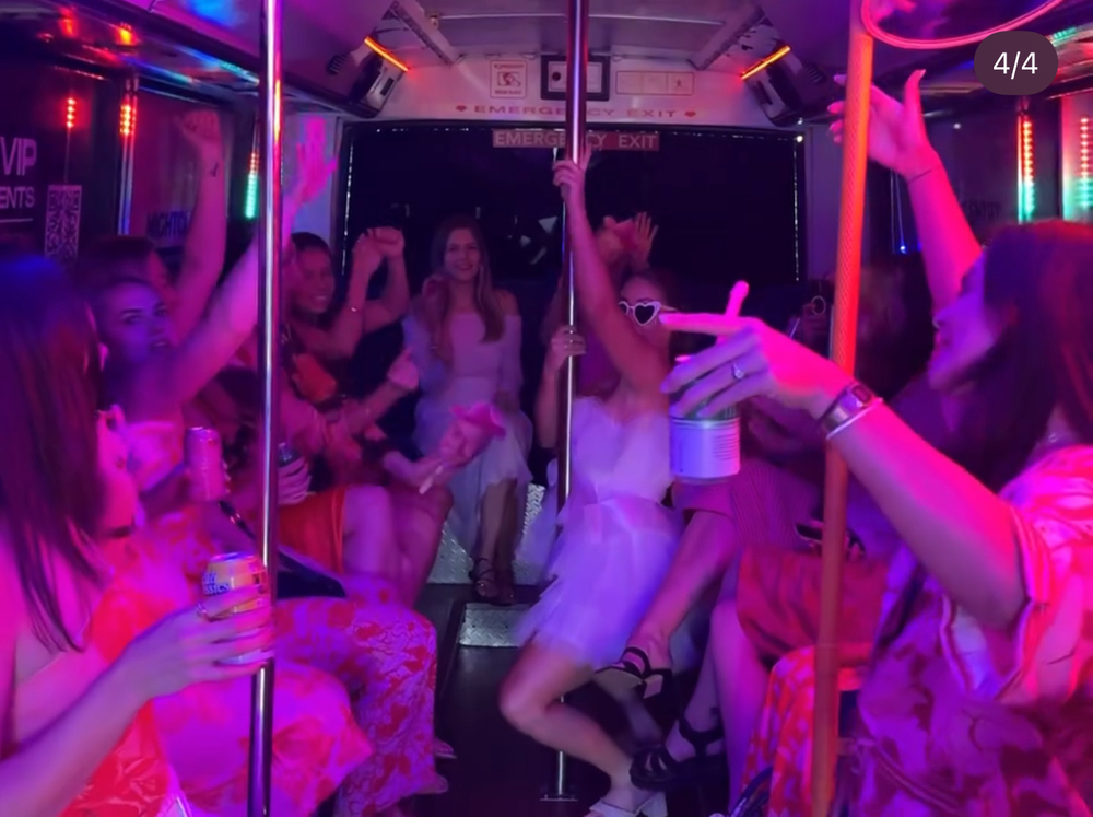 A hens group dancing and singing inside a party bus lit pink and purple, one friend in heart-shaped sunglasses by the pole