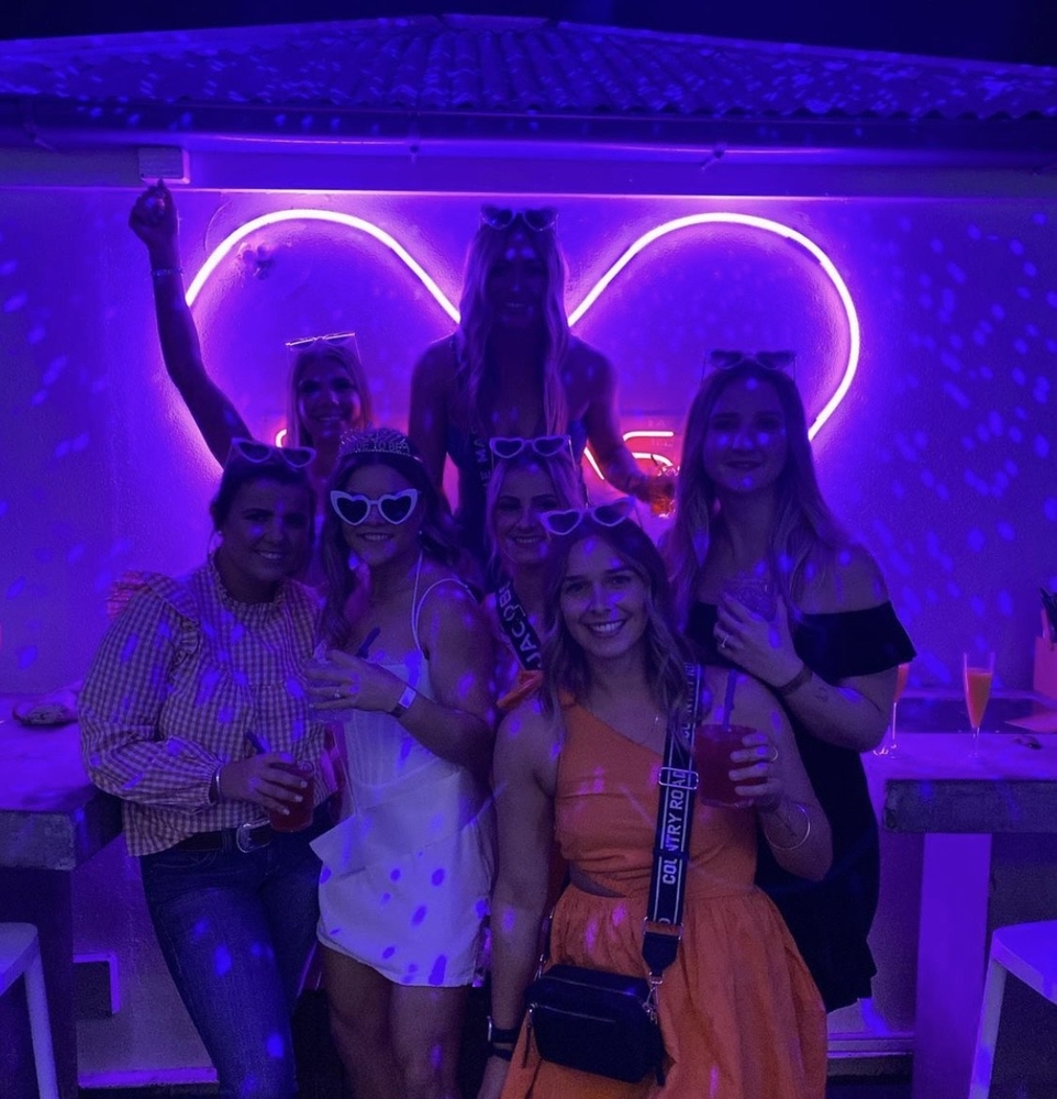 A hens group in heart-shaped sunglasses posing under a glowing neon heart in a purple-lit bar