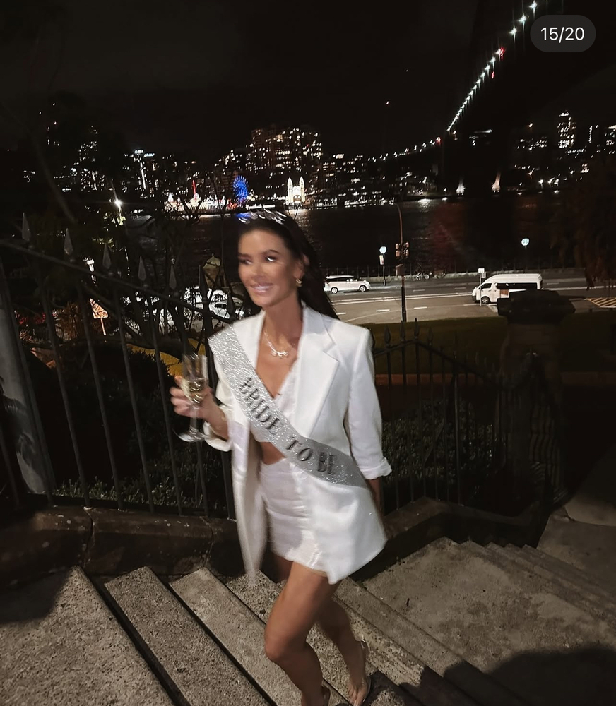 Britt in a white blazer and Bride to Be sash on the harbour steps at night, glass in hand