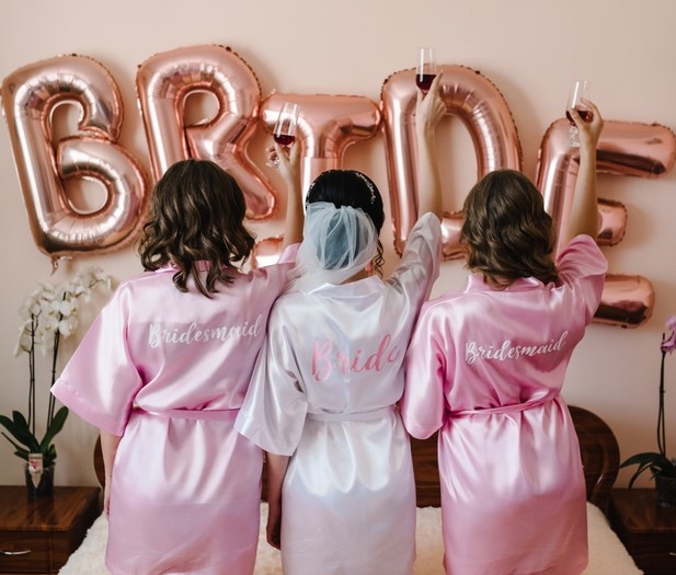 The bride and two friends in pink satin robes facing a wall of rose gold BRIDE balloons at their hens weekend accommodation