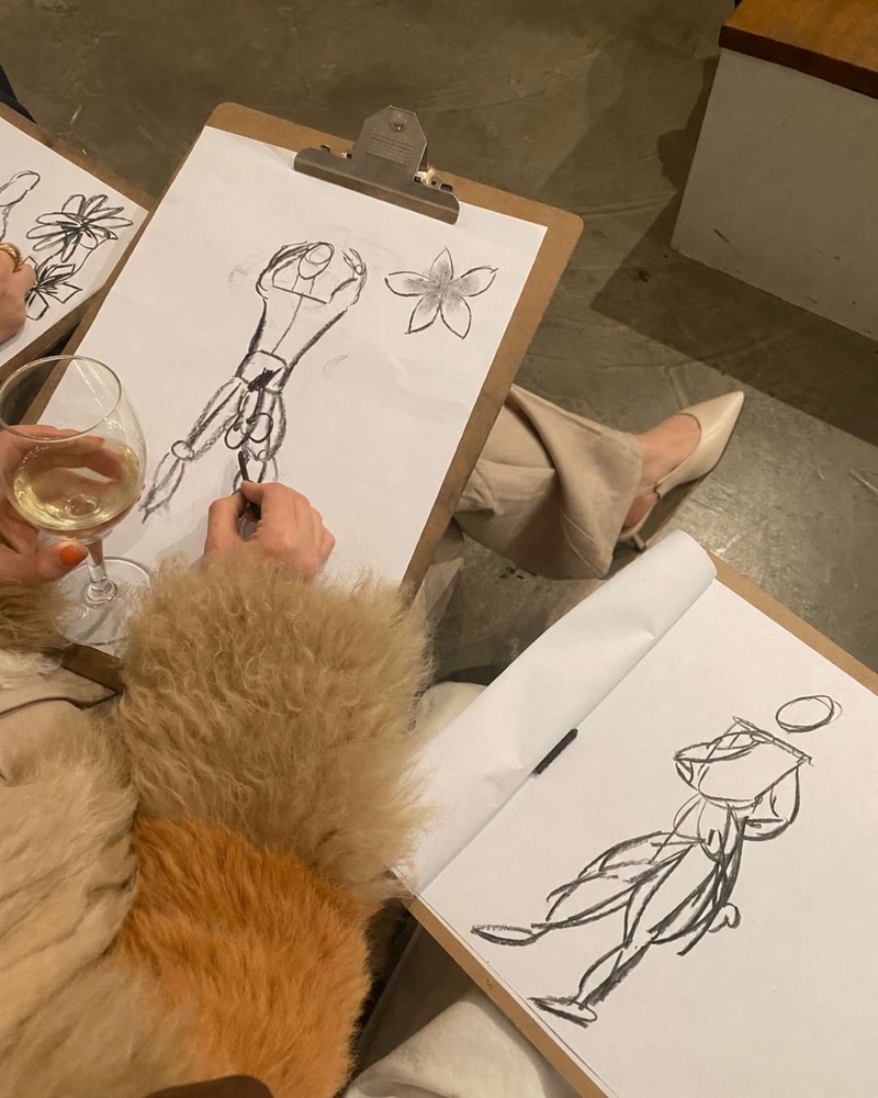 Life drawing sketches on a clipboard beside a glass of white wine