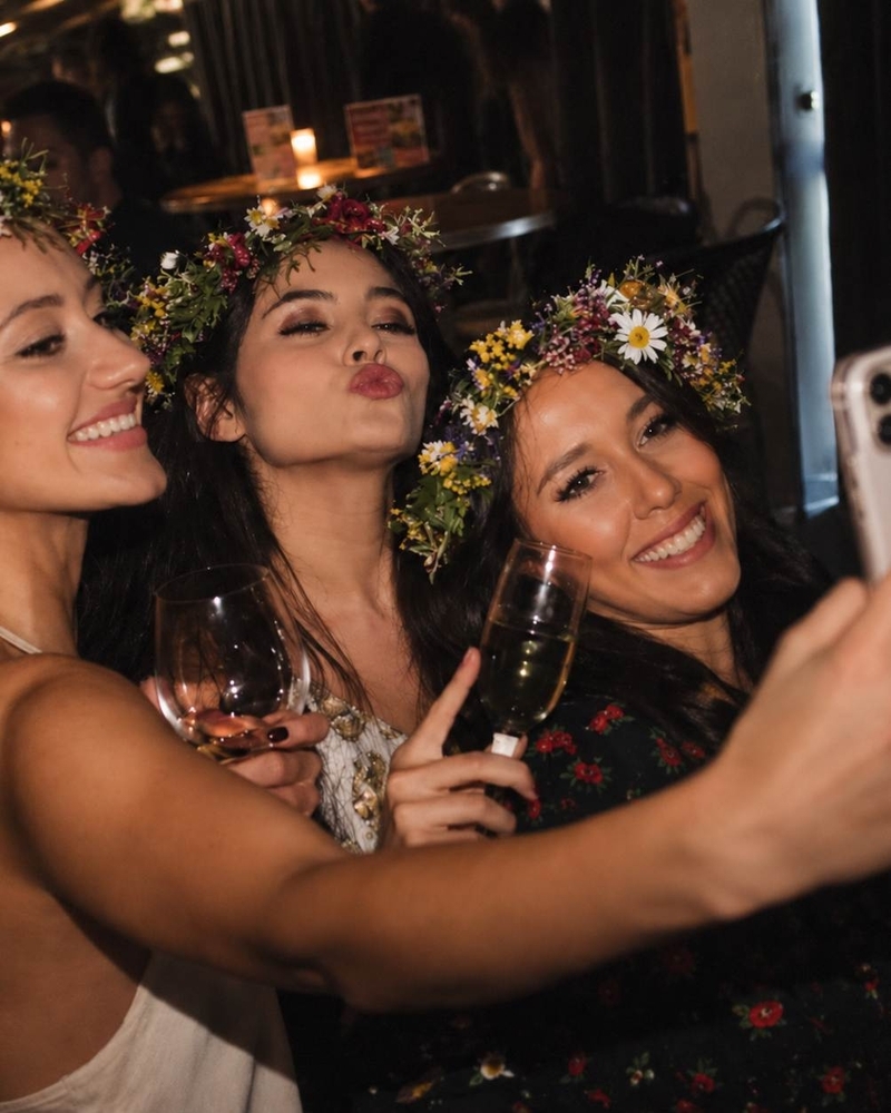 Three friends in flower crowns squeezing into a selfie with glasses of bubbles