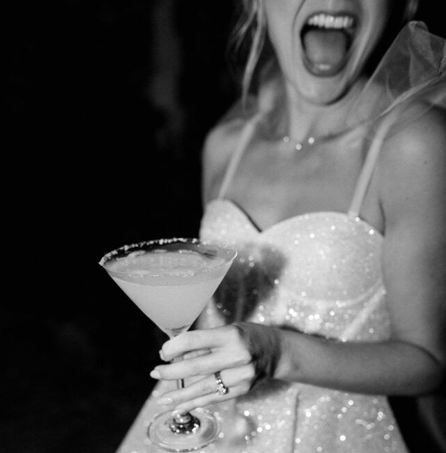 Black and white photo of a bride to be in a veil, laughing with a martini