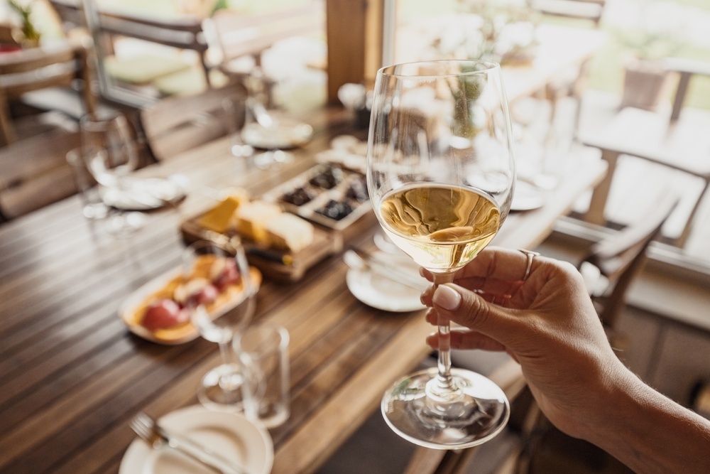 A glass of white wine raised over a sunny outdoor table set for lunch