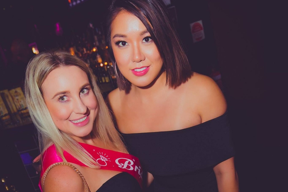 A smiling bride in a pink sash cheek to cheek with a friend in a dim cocktail lounge